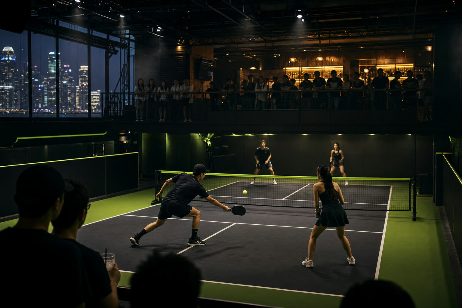 Spectators watching a doubles match from a mezzanine bar above a floodlit indoor court